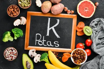 Tablica z napisem "K Potas" otoczona owocami i warzywami bogatymi w potas, jak banany, awokado, pomidory, ziemniaki, brokuły, rodzynki, morele, kiwi, migdały, pestki dyni. Pomaga zapobiegać hipokaliemii.