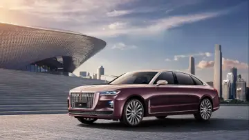 Elegancki, bordowy Hongqi H9 z chromowanymi detalami stoi przed nowoczesnym budynkiem o falistej fasadzie, w tle miejski krajobraz.