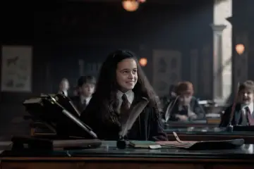 Uśmiechnięta uczennica w szacie Hogwartu, część obsady "Harry Potter i Kamień Filozoficzny", siedzi przy ławce z piórem i kałamarzem.