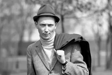 Mężczyzna w kapeluszu i golfie, z marynarką na ramieniu, jakby przygotowywał się do występu z piosenką "Daria Zawiałow jeszcze w zielone gramy".