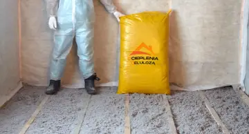 Osoba w kombinezonie stoi obok worka z materiałem do ocieplenia celulozą. Podłoga pokryta jest już warstwą izolacji.