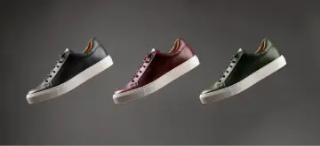 Sneakers marche famose in pelle nera, bordeaux e verde, con suola bianca, fluttuano su sfondo grigio.