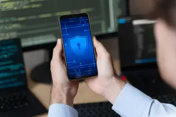 Ręce trzymające smartfon z ikoną tarczy i klucza. W tle monitory z kodem. Szukasz sposobu, jak usunąć blokadę rodzicielską w telefonie?