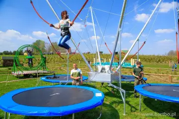 Dziewczyna skacze na eurobungee, unosząc się wysoko nad trampolinami. W tle widać inne atrakcje i ludzi.