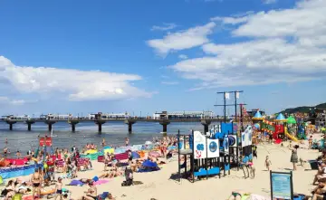 Tłum ludzi cieszy się słońcem i zabawą na plaży w Międzyzdrojach. W tle widać molo, a na piasku kolorowy plac zabaw.