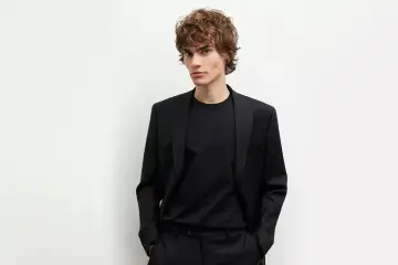 Młody mężczyzna w czarnej marynarce i czarnych spodniach, z rękami w kieszeniach, prezentuje elegancki styl.