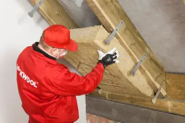Pracownik w czerwonym kombinezonie montuje wełnę mineralną, wykonując ocieplenie murłaty od wewnątrz.