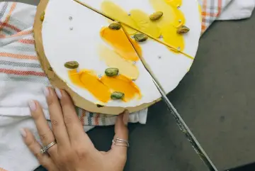 Dłoń kroi tort na 30 osób. Biały krem, żółte dekoracje i pistacje.
