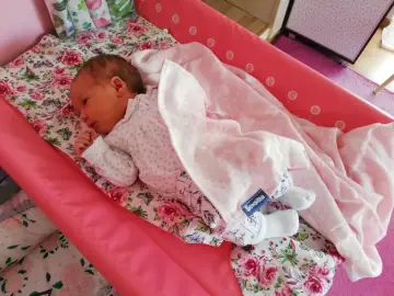 Nowe dziecko śpi otulone w pieluchę tetrową na różowej macie. Pokazano, jak zawijać dziecko w pieluchę tetrową, by czuło się bezpiecznie.