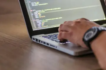 Kodowanie strony internetowej, widać fragment kodu HTML z listą nieuporządkowaną i rękę piszącą na klawiaturze.