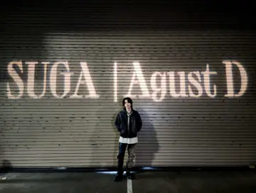 Agust D, czyli Suga z BTS, stoi przed rolowaną bramą z napisem "SUGA | Agust D" wyświetlonym na niej.