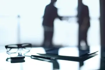 Okulary, długopis i tablet na biurku. W tle sylwetki dwóch mężczyzn, być może omawiających kwestię opłaty skarbowej.