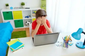 Chłopiec zamyka twarz w dłoniach, martwiąc się, czy można nie dostać się do żadnej szkoły. Na biurku laptop, zeszyty i lampka.