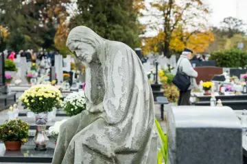 Kamienna figura pochyla się nad grobami. W tle cmentarz, gdzie można szukać bliskich za pomocą ogólnopolska wyszukiwarka osób pochowanych.