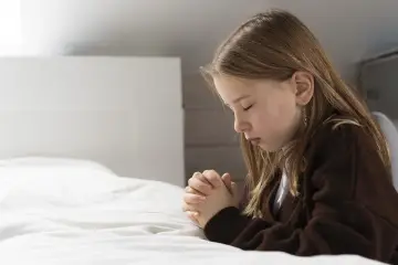Dziewczynka z zamkniętymi oczami, składając dłonie, wykonuje akt żalu dla dzieci. Modli się na białej pościeli.