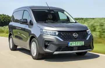 Szary Nissan Townstar jedzie po drodze, otoczony zielonymi polami. Kierowca w okularach prowadzi pojazd.