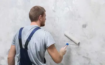 Mężczyzna w ogrodniczkach przygotowuje ściany do malowania, nakładając biały grunt wałkiem.