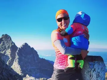 Justyna Kowalczyk z dzieckiem na szczycie górskim, uśmiechnięta, w otoczeniu skalistych szczytów i błękitnego nieba.