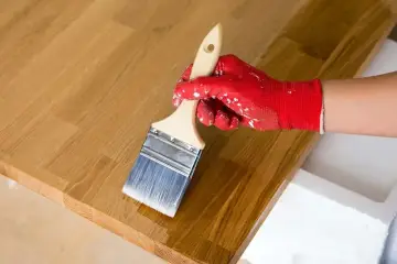 Application d'huile de lin sur un plateau en bois de douglas. La main gantée de rouge applique le produit avec un pinceau.