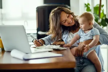 Uśmiechnięta mama z dzieckiem na rękach wypełnia dokumenty. Może to być wniosek o subwencję na dziecko z orzeczeniem o potrzebie kształcenia specjalnego.