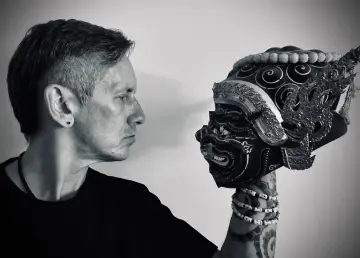 Mężczyzna w czarnym t-shircie patrzy na tradycyjną maskę. Czerń koloru podkreśla tajemniczość sceny.