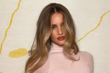 Kobieta z pięknymi, falowanymi włosami w odcieniu honey blonde, w różowym golfie, na tle wzorzystej ściany.
