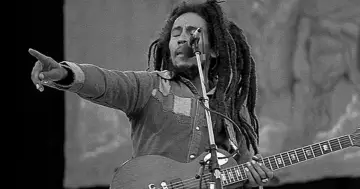 Bob Marley, z charakterystycznymi dredami, śpiewa do mikrofonu, grając na gitarze. Jego muzyka inspirowała wielu, w tym jego liczne bob marley dzieci.