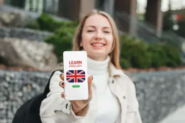 Uśmiechnięta dziewczyna trzyma telefon z napisem "LEARN ENGLISH" i flagą Wielkiej Brytanii. To darmowa aplikacja do nauki angielskiego.