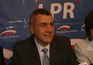 Mężczyzna w garniturze, z niebieskim krawatem, na tle banera z napisem "LPR" i hasłami "Mądra edukacja" oraz "Bezpieczna rodzina".