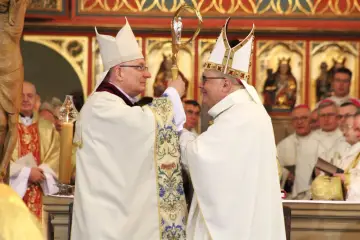 Dwóch biskupów w białych szatach i mitrach podczas ingresu do katedry. Jeden przekazuje drugiemu pastorał.