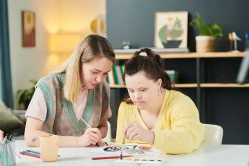 Dwie kobiety malują, jedna z zespołem Downa. Wspólne zajęcia artystyczne to element oligofrenopedagogiki, rozwijający kreatywność i umiejętności.