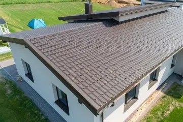Nowy dom z brązowym, falistym dachem z miedzi. W oddali widać niebieski namiot na zielonym trawniku.