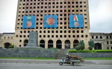 Zniszczony budynek w Abchazji z wielkimi banerami z medalami. Na pierwszym planie mężczyzna na skuterze z przyczepą pełną drewna.