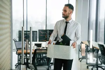 Mężczyzna w białej koszuli i krawacie wychodzi z biura z pudełkiem osobistych rzeczy. To jedna z przyczyn bezrobocia.