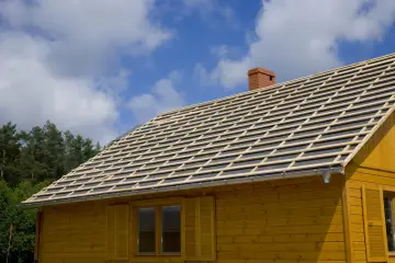 Drewniany dom z nowym dachem w trakcie budowy. Widoczne łaty pod blachodachówkę, komin i okna z okiennicami.