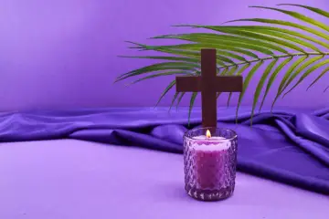 Drewniany krzyż, płonąca świeca i gałązka palmy na fioletowym tle. Symbolika postu i refleksji.