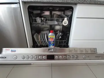 Zmywarka Bosch z otwartymi drzwiczkami, w środku naczynia i butelka środka do czyszczenia zmywarki Kret.