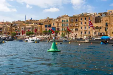 Malta w sierpniu: port z łodziami i kamiennymi budynkami, ozdobiony flagami.