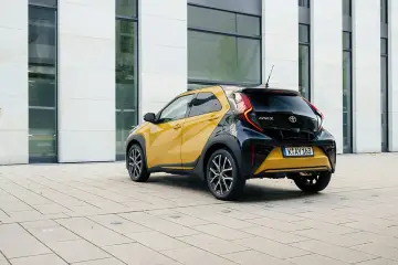 Żółto-czarny Toyota Aygo X Hybrid zaparkowany na placu przed nowoczesnym budynkiem.