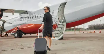 Uśmiechnięta stewardessa z walizką przy samolocie. Czas na boarding, czyli wejście na pokład.