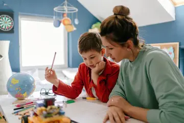 Chłopiec z ołówkiem w ręku uczy się z mamą. Czy nauczanie domowe jest płatne? Na biurku globus i kredki.