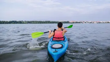 Mężczyzna w kamizelce ratunkowej wiosłuje kajakiem po rzece Reda. W tle widać miasto i zieleń.