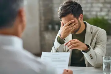 Mężczyzna z ręką na twarzy, siedzący przy biurku z dokumentami i okularami.