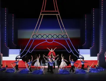 Tancerze baletowi na scenie z podświetloną Wieżą Eiffla. W tle czerwono-niebieska scenografia, przypominająca francuską flagę. To piękna aria, która porusza serca.