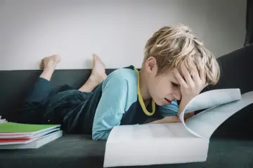 Chłopiec z ręką na czole, sfrustrowany nauką. Stos zeszytów obok. To trudny ii etap edukacyjny.