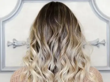 Włosy z efektem ombre blond, falowane, z ciemniejszymi końcówkami przechodzącymi w jasny blond.