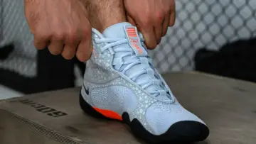 Ręce zapinają jasnoniebieskie buty sportowe z pomarańczowymi akcentami, przypominające styl Batistini.
