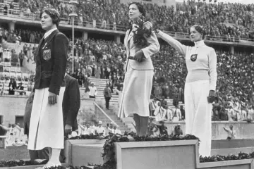 Trzy medalistki na podium, wśród nich triumfatorka z wieńcem laurowym. Scena przypomina pierwsze igrzyska olimpijskie.