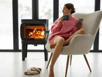 Kobieta w różowym swetrze pije kawę, relaksując się przy ogniu w kominku. Zastanawia się, gdzie kupić brykiet na zimowe wieczory.