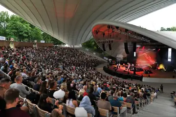 Koncert w jednym z polskich amfiteatrów. Tłum widzów siedzi na widowni, podziwiając występ zespołu na oświetlonej scenie.